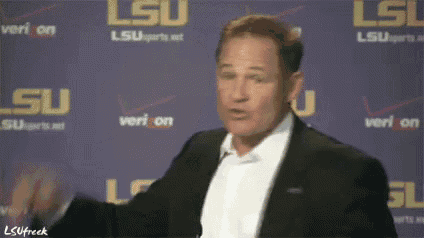 a man in a suit stands in front of a purple wall with lsu written on it