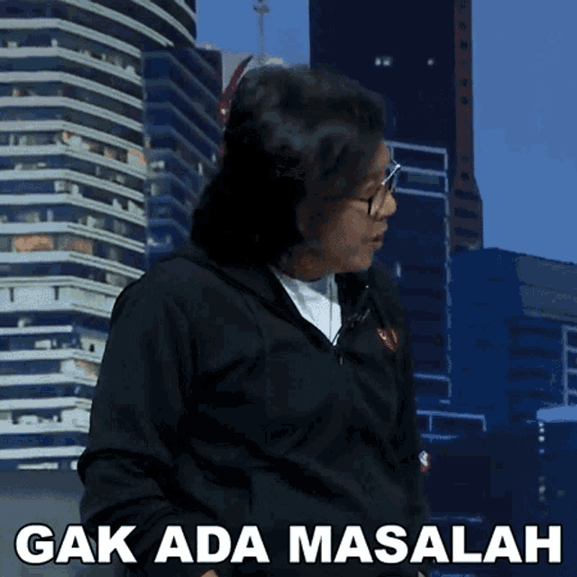 a man in a black jacket stands in front of a city skyline with the words " gak ada masalah " on the bottom