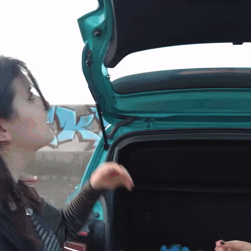 a woman opens the trunk of a car with graffiti in the background
