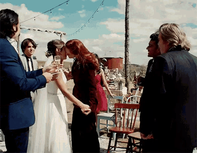 a group of people are standing around a bride and groom kissing while holding glasses of champagne .