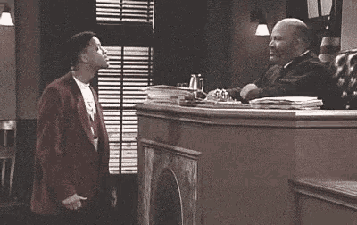 a man in a red suit is standing in front of a judge in a courtroom talking to him .