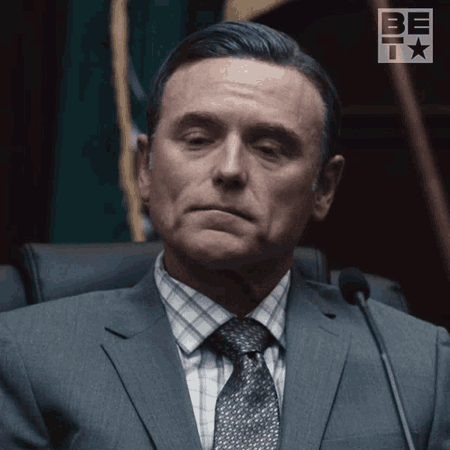 a man in a suit and tie is sitting in front of a microphone with the word be on the bottom