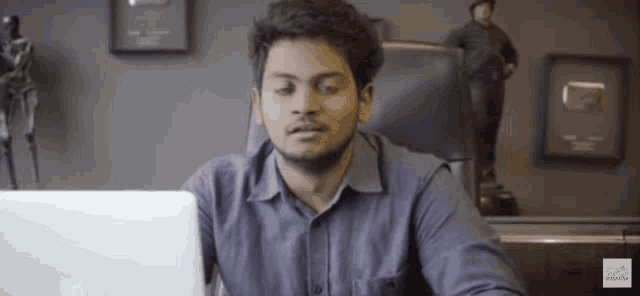 a man in a blue shirt is sitting in front of a laptop computer .