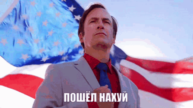 a man in a suit and tie stands in front of an american flag with his hands on his chest