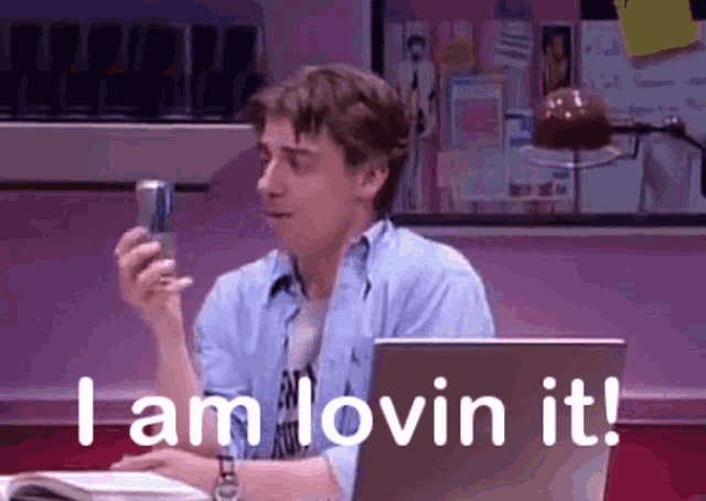 a man is sitting in front of a laptop holding a cell phone and saying i am loving it .