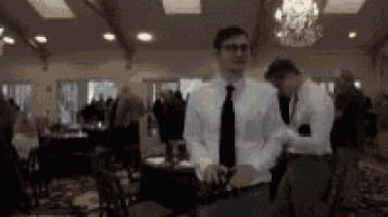 a man in a white shirt and black tie is standing in a room with people .