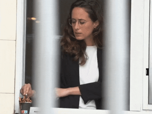 a woman wearing glasses looks out of a window with a cup of coffee in front of her