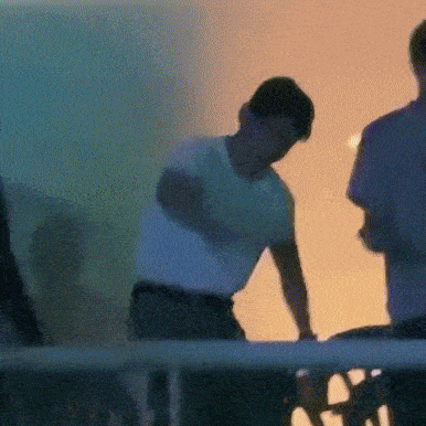 a man in a white shirt is standing in front of a fence with a sunset in the background .