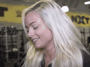 a blonde woman is smiling in front of a yellow sign that says nxt
