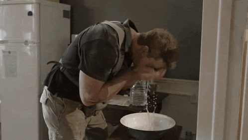 a man washing his face with a bowl of water in front of a refrigerator