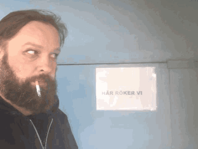 a man with a beard is smoking a cigarette in front of a sign that says " har röker vi "