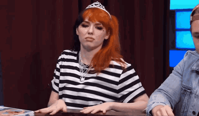 a woman with red hair and a tiara is sitting at a table with her hands on the table .