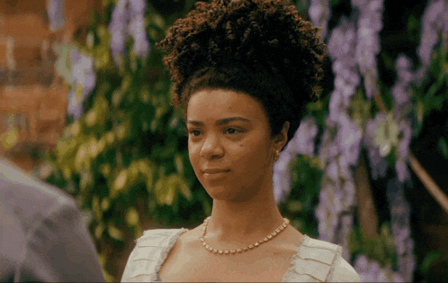 a woman with curly hair and a pearl necklace stands in front of purple flowers