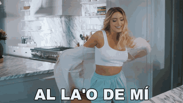 a woman in a white top and blue shorts is dancing in a kitchen with the words al la ' o de mi behind her