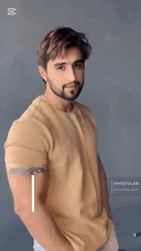a man with a beard is wearing a tan t-shirt with his hands in his pockets
