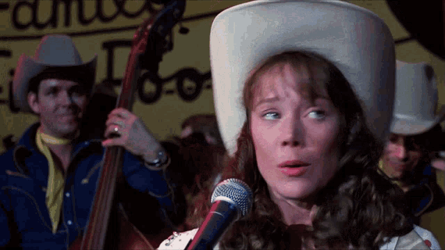 a woman in a cowboy hat sings into a microphone in front of a man playing a bass