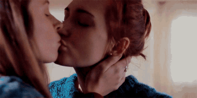 two women are kissing each other in a room . one of the women is wearing a blue sweater .