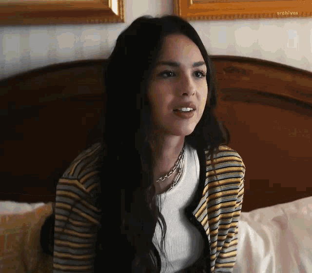 a woman is sitting on a bed wearing a striped cardigan and a white shirt .