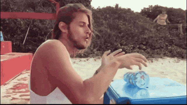 a man is sitting at a table on the beach smoking a cigarette and drinking from a plastic bottle .