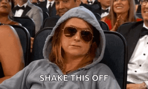 a woman wearing a hoodie and sunglasses is sitting in a crowd and says shake this off