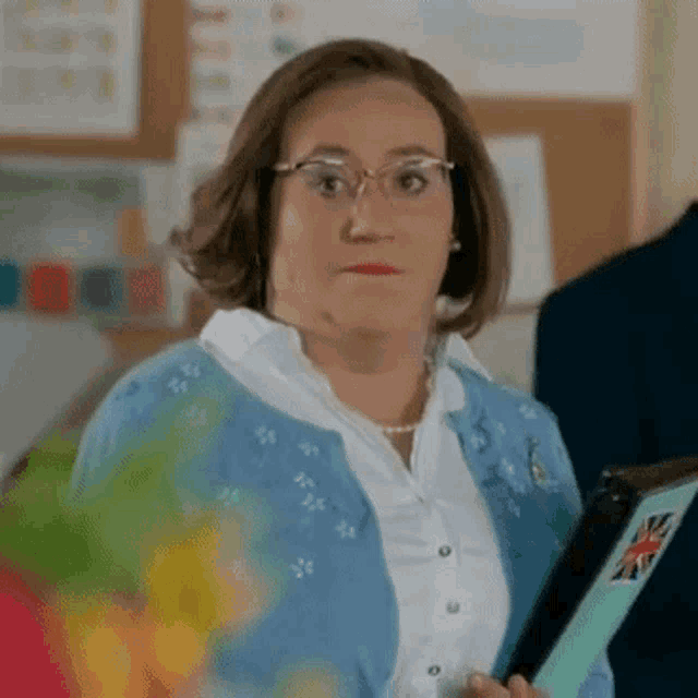 a woman wearing glasses and a blue cardigan is holding a clipboard .
