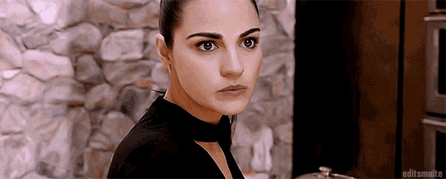 a woman in a black top is standing in front of a stone wall looking at the camera