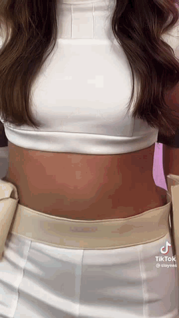a woman is wearing a white crop top and white pants with a tan belt around her waist .