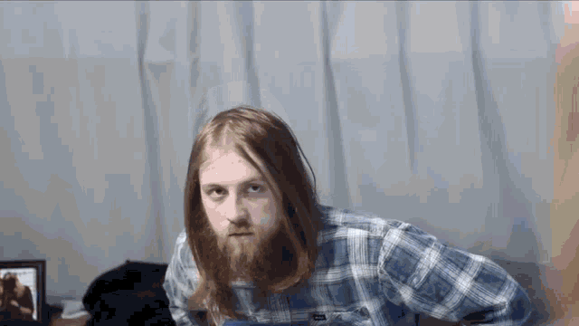 a man with long hair and a beard wearing a plaid shirt is sitting on a bed .