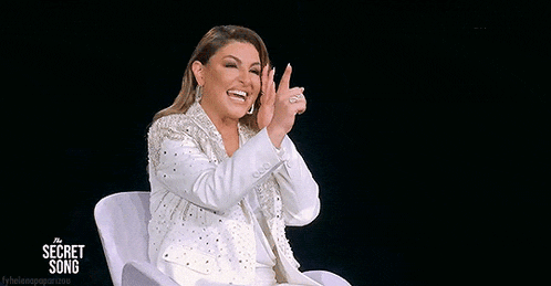 a woman in a white suit is sitting in a chair clapping her hands .
