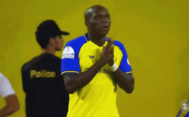 a man in a yellow and blue shirt is clapping in front of a man in a black shirt that says police