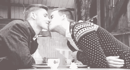 two men are kissing each other in a black and white photo while sitting at a table .