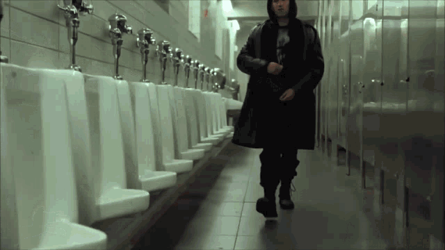 a man in a hooded coat walks through a row of urinals in a public restroom