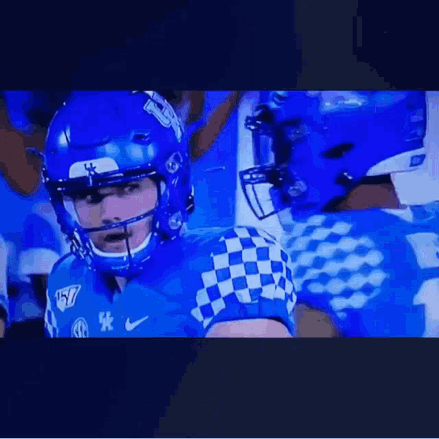 a football player wearing a blue helmet and a blue and white checkered jersey is standing in front of a crowd .