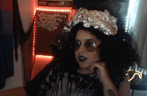 a woman wearing sunglasses and a tin foil crown stands in front of a happy halloween sign