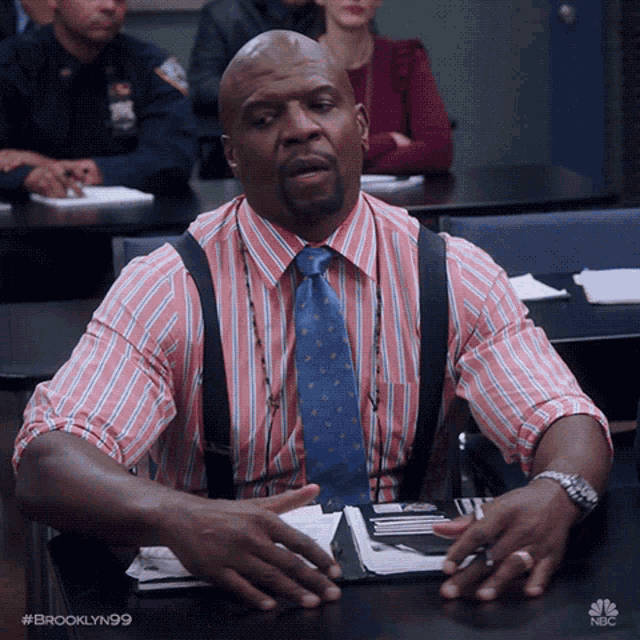 a man in a pink and blue striped shirt and tie sits at a desk with his hands folded in front of his face