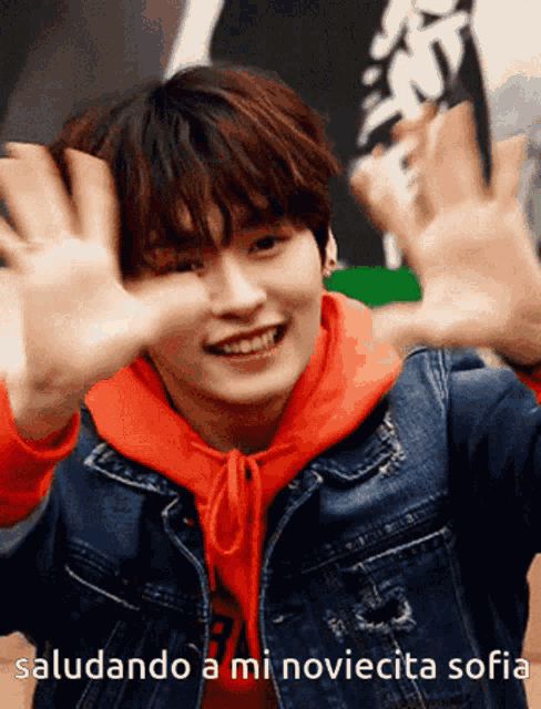 a young man wearing a red hoodie and a denim jacket is smiling and waving his hands
