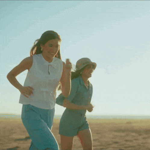 two women are running on the beach and one is wearing a straw hat