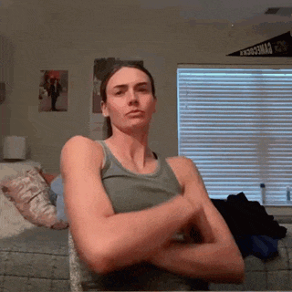 a woman in a tank top is standing with her arms crossed in front of a window with blinds ..