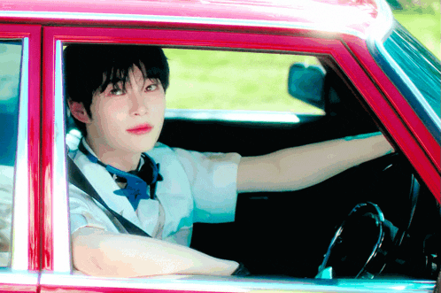 a young man is driving a red car and looking out the window