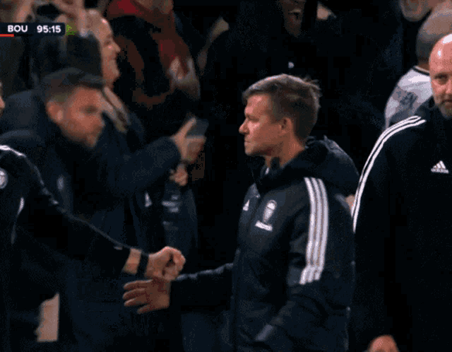 a man in a black adidas jacket is shaking hands with another man in the stands