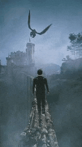 a man is standing on top of a rocky cliff looking at a dragon flying overhead .