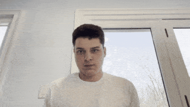 a man in a white shirt holds his hand to his head in front of a window