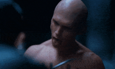 a bald man with a tattoo on his chest looks at another man