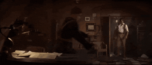 two men are standing in a dark room with papers on the floor and a lamp