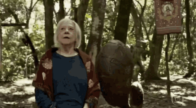 an elderly woman is standing in the middle of a forest with a statue in the background .