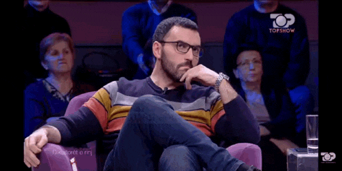 a man with glasses is sitting in a chair in front of a crowd of people .