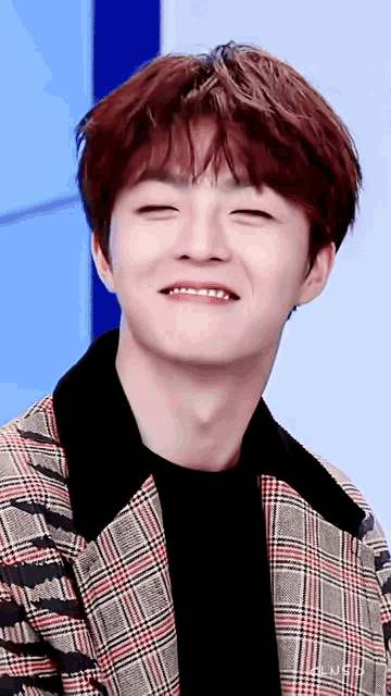 a young man wearing a plaid jacket and a black shirt smiles with his eyes closed