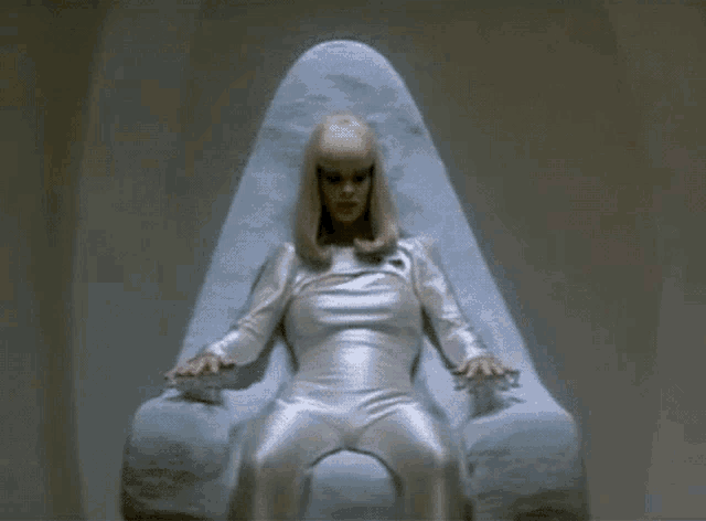 a woman in a gold bodysuit is sitting on a throne with her hands on the buttons .