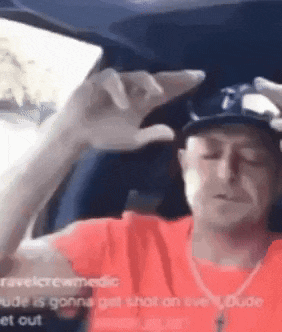 a man in a red shirt and hat is sitting in a car holding a cell phone to his head .