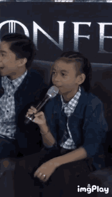 two young boys are sitting on a couch and one is holding a microphone while the other looks on